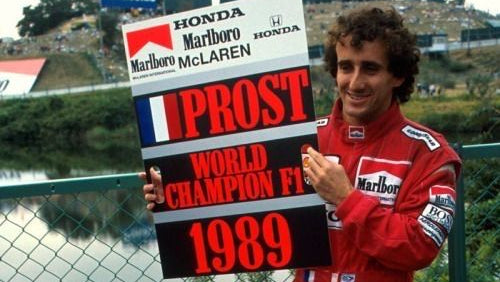 Alain Prost  in red racing suit holding a 'Prost World Champion 1989' sign with a world map umbrella in the foreground.